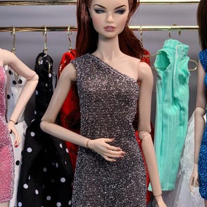 May include: A doll wearing a silver sparkly one-shoulder dress. The doll has long, dark brown hair styled in a bouffant. The doll is standing in front of a rack of clothing.