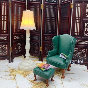 Pu&ograve; includere: Una poltrona a orecchie verde in miniatura con un poggiapiedi e una lampada bianca con un paralume bianco. La poltrona e il poggiapiedi sono entrambi rivestiti in un tessuto verde scuro. La lampada &egrave; su una base bianca e ha un paralume bianco. La poltrona e il poggiapiedi sono davanti a uno schermo di legno.