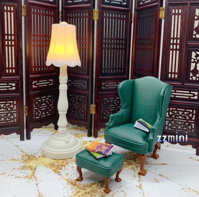 Pu&ograve; includere: Una poltrona a orecchie verde in miniatura con un poggiapiedi e una lampada bianca con un paralume bianco. La poltrona e il poggiapiedi sono entrambi rivestiti in un tessuto verde scuro. La lampada &egrave; su una base bianca e ha un paralume bianco. La poltrona e il poggiapiedi sono davanti a uno schermo di legno.