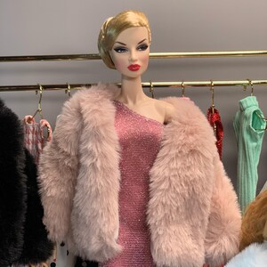 May include: A fashion doll wearing a pink faux fur jacket over a sparkly pink dress. The doll has blonde hair styled in a bun and red lipstick. The doll is standing in front of a rack of clothing.