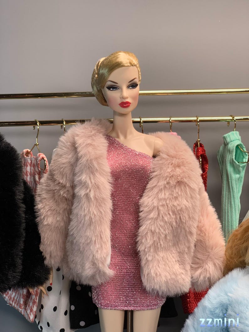 May include: A fashion doll wearing a pink faux fur jacket over a sparkly pink dress. The doll has blonde hair styled in a bun and red lipstick. The doll is standing in front of a rack of clothing.