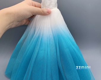 Peut inclure: Une robe en tulle à dégradé de blanc à bleu avec un corsage en dentelle. La robe est conçue pour une poupée et a une jupe ample.