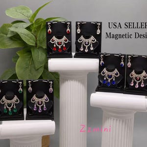 May include: Six sets of jewelry displayed on black stands, each featuring a necklace and matching earrings with teardrop-shaped crystals in red, clear, blue, green, purple, and pink. The text USA SELLER Magnetic Design is visible.