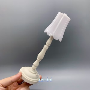 Pu&ograve; includere: Una lampada da tavolo bianca con un paralume bianco a balze. La lampada ha una base decorativa con una colonna scanalata.