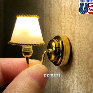 Könnte beinhalten: Eine goldene Miniatur-Wandleuchte mit einem weißen Stofflampenschirm. Die Leuchte hat einen dekorativen goldenen Rand um den Lampenschirm und einen runden Sockel.