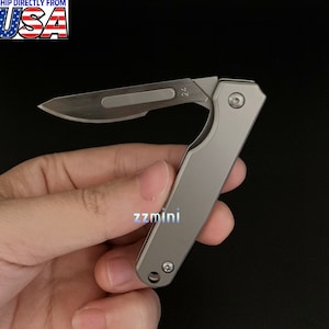 May include: A small, silver folding knife with a partially open blade. The blade has a sharp point and a small, rectangular cutout. The handle is smooth and rectangular. The text "24" is visible on the blade. The text "zzmini" is visible on the handle.
