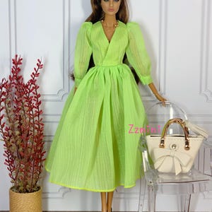 May include: A lime green dress with a V-neckline and long sleeves. The dress has a gathered skirt and a fitted bodice. The doll is wearing a gold necklace and holding a white handbag.