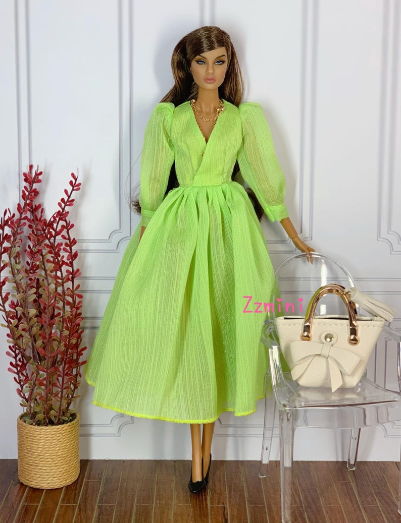 May include: A lime green dress with a V-neckline and long sleeves. The dress has a gathered skirt and a fitted bodice. The doll is wearing a gold necklace and holding a white handbag.