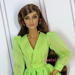 May include: A doll wearing a lime green dress with a ruffled skirt and long sleeves. The doll has long brown hair, blue eyes, and is wearing gold jewelry and red earrings.