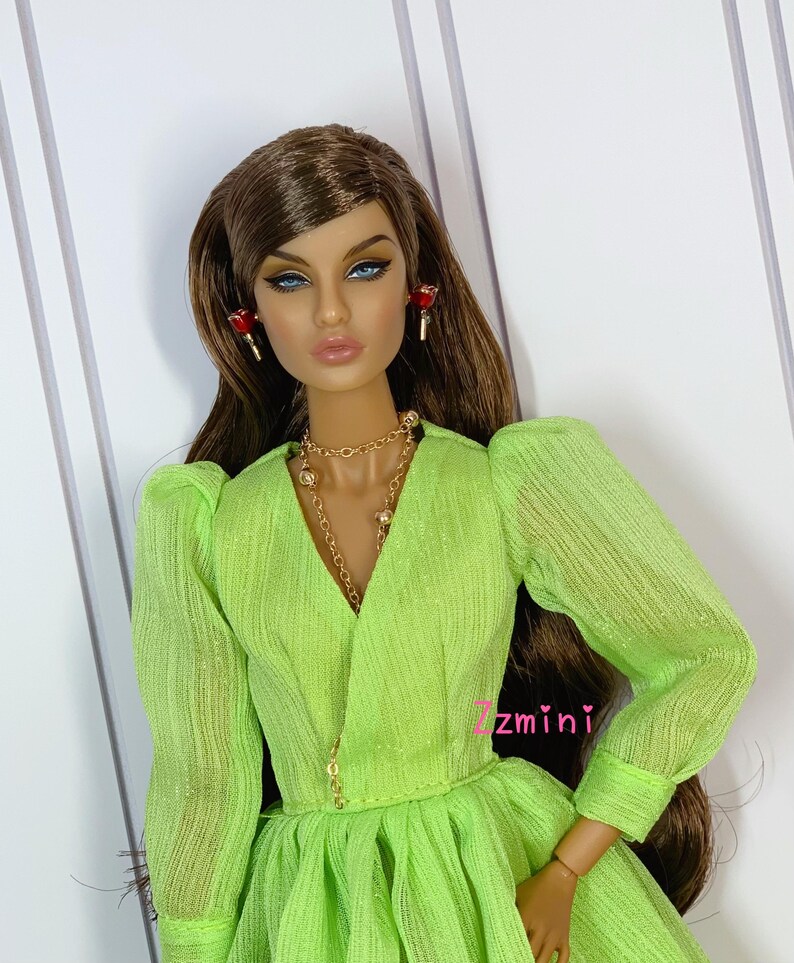 May include: A doll wearing a lime green dress with a ruffled skirt and long sleeves. The doll has long brown hair, blue eyes, and is wearing gold jewelry and red earrings.