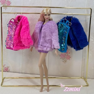 May include: A fashion doll with blonde hair stands in front of a gold metal clothing rack. The rack displays a pink sequined top with a pink faux fur jacket, and a blue sequined top with a blue faux fur jacket. The doll wears a purple faux fur jacket.
