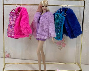 May include: A fashion doll with blonde hair stands in front of a gold metal clothing rack. The rack displays a pink sequined top with a pink faux fur jacket, and a blue sequined top with a blue faux fur jacket. The doll wears a purple faux fur jacket.