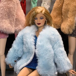May include: A doll wearing a light blue faux fur coat over a sparkly blue dress. The doll has blonde hair and is sitting on a clear acrylic chair.