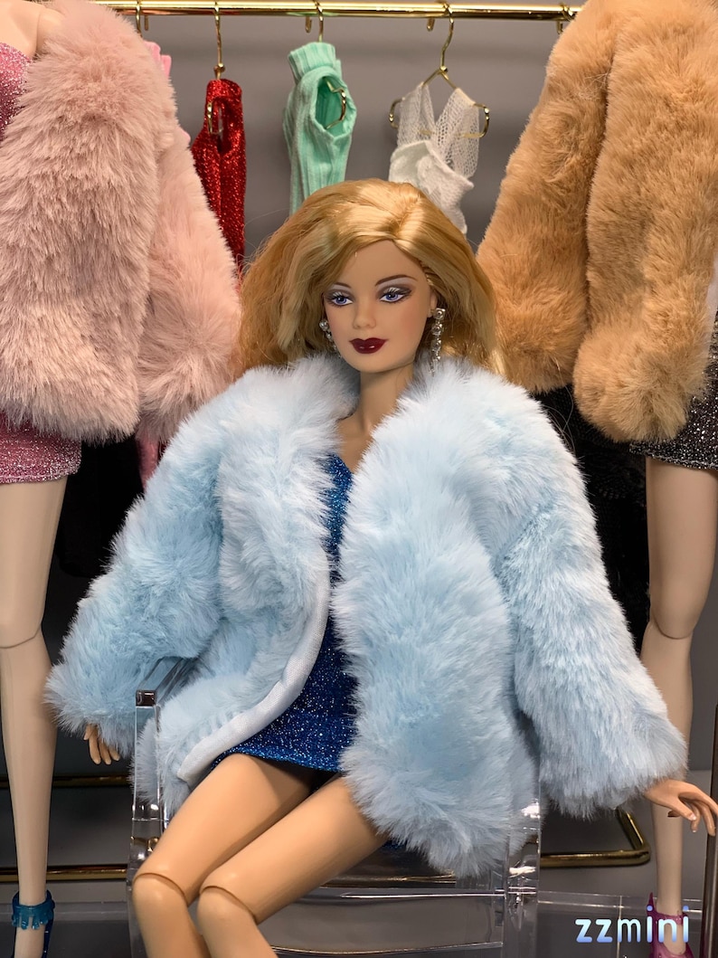 May include: A doll wearing a light blue faux fur coat over a sparkly blue dress. The doll has blonde hair and is sitting on a clear acrylic chair.