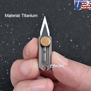 May include: A small titanium pocket knife with a silver blade and a brass button. The knife is held in a hand.