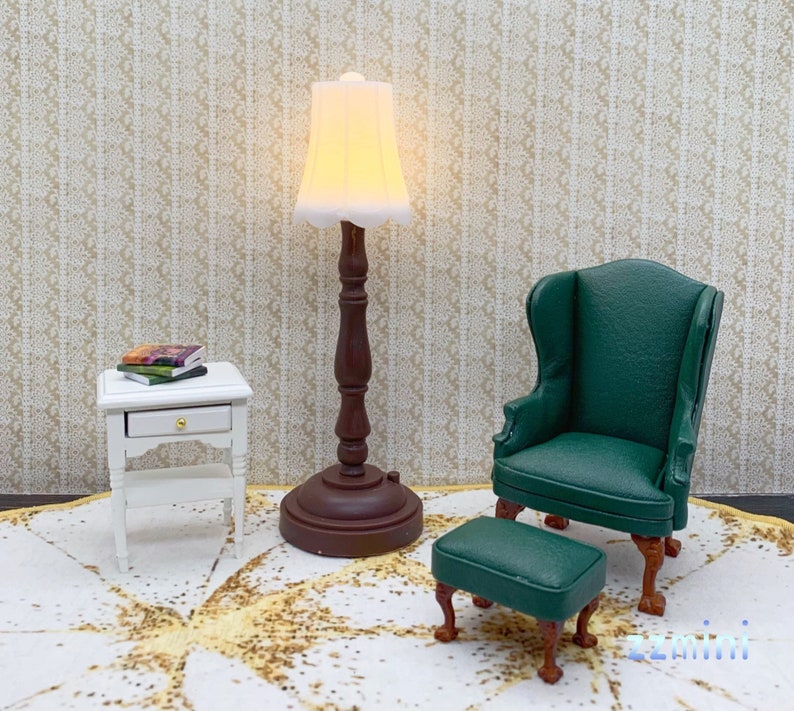 Pu&ograve; includere: Una lampada bianca in miniatura con un paralume color crema si trova su un tappeto bianco accanto a un tavolino bianco con un cassetto. Una poltrona verde con un poggiapiedi abbinato si trova sul tappeto.