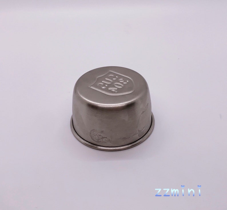 May include: A silver metal cup with a rounded top and a raised shield design on the top. The shield has the text "304 SUS" embossed on it.
