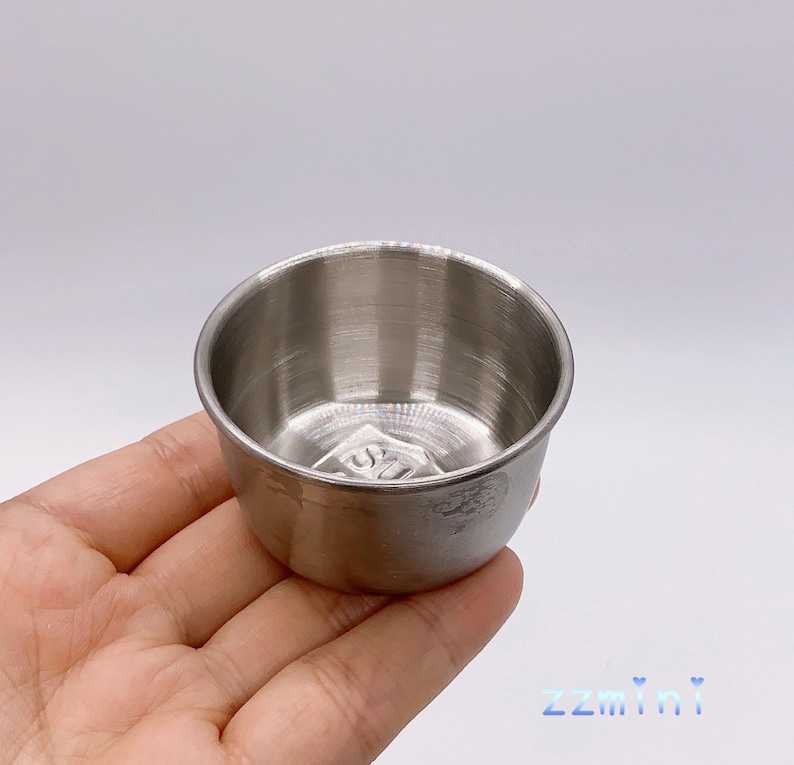 May include: A small, silver, stainless steel cup with a rounded bottom. The cup is about 2 inches in diameter and 1.5 inches tall.