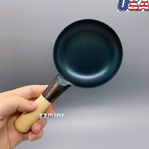 May include: A small, black, cast iron frying pan with a wooden handle. The pan is about 6 inches in diameter.