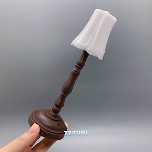 Pu&ograve; includere: Una lampada da tavolo in legno marrone in miniatura con un paralume in tessuto bianco. La lampada &egrave; alta circa 15 cm.