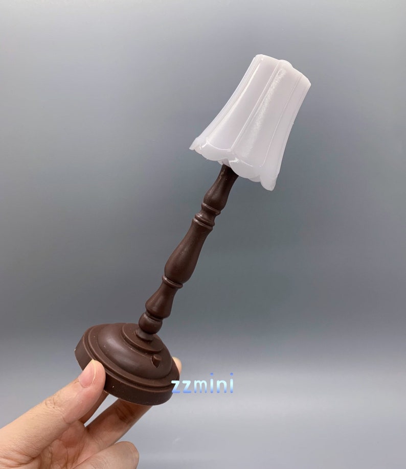 Pu&ograve; includere: Una lampada da tavolo in legno marrone in miniatura con un paralume in tessuto bianco. La lampada &egrave; alta circa 15 cm.