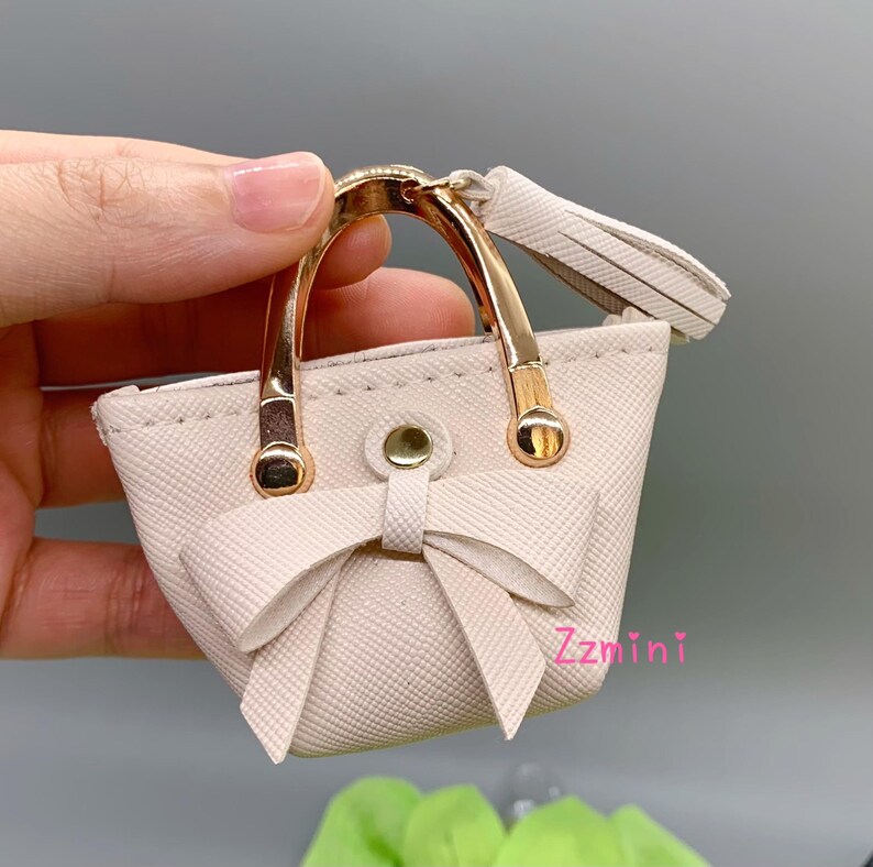 May include: A small white faux leather handbag with gold-tone hardware and a bow detail. The bag has a top handle and a tassel.