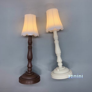 Pu&ograve; includere: Due lampade da tavolo in miniatura con paralumi bianchi. La lampada a sinistra ha una base marrone e la lampada a destra ha una base bianca. Le lampade sono accese e i paralumi sono illuminati.