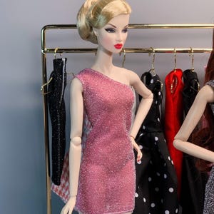 May include: A pink sparkly one-shoulder dress for a fashion doll. The dress is displayed on a doll stand in front of a rack of clothing.