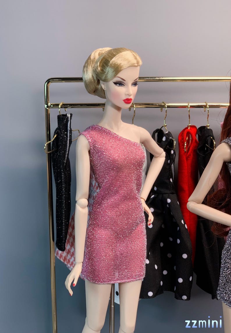 May include: A pink sparkly one-shoulder dress for a fashion doll. The dress is displayed on a doll stand in front of a rack of clothing.