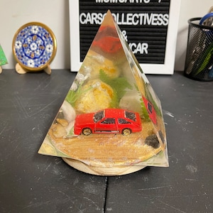 May include: A red toy car encased in a clear pyramid-shaped resin block with a wooden base. The resin block contains small plants and rocks.