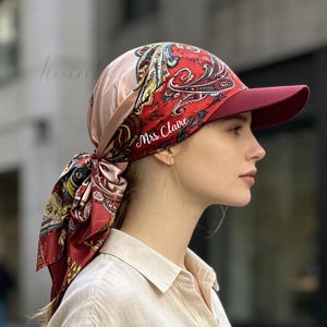 May include: A baseball cap featuring a red, gold, and black paisley patterned fabric that extends into a tied scarf at the back. The cap has a solid dark red brim and the text Mrs Claire embroidered in white script on the side of the crown.