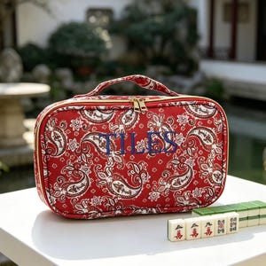 Könnte beinhalten: Eine rechteckige rote Aufbewahrungstasche mit Reißverschluss, weiß-schwarzem Paisleymuster und dem in Blau gestickten Wort TILES auf der Vorderseite. Die Tasche hat einen Tragegriff und goldfarbene Reißverschlüsse sowie eine Reihe Mahjong-Steine.