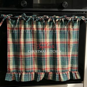 Pode incluir: Uma capa de forno em tecido xadrez verde, vermelho e amarelo com uma bainha franzida e laços para prender. O centro está bordado com o texto em vermelho "Sophia's" acima do texto em branco "CHRISTMAS KITCHEN".