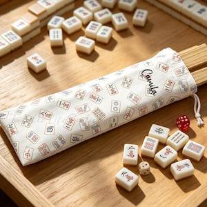 May include: A white fabric drawstring pouch featuring a repeating pattern of Mahjong tiles and the name Camila printed in black script. The pouch contains wooden racks and is surrounded by scattered ivory-coloured Mahjong tiles and two red dice.