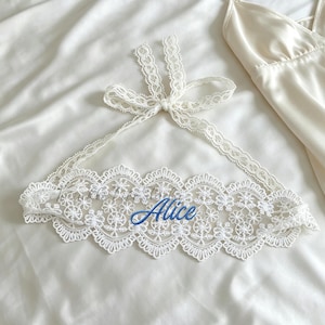 Op de afbeelding: Een witte kanten haltertop met een bloemmotief en een striksluiting in de nek, vergezeld van een bijpassende witte satijnen slipdress met dunne spaghettibandjes. Het woord Alice is in blauw schrift geborduurd in het midden van de kanten top.