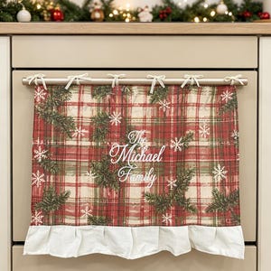 May include: A rectangular oven towel featuring a red, green, and cream plaid pattern with printed pine branches and white snowflakes. The centre displays the white text The Michael Family. It has a white ruffled bottom edge and tie-top loops.
