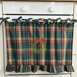 May include: A green, red and tan tartan cafe curtain featuring a ruffled bottom edge and tie-top tabs. The centre displays an embroidered design of a red bow atop a green wreath encircling the red cursive letter N on the fabric surface.
