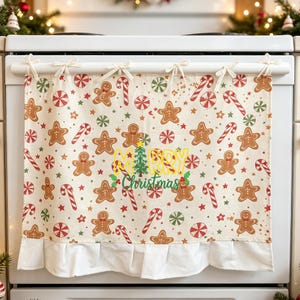 May include: A cream-coloured oven door curtain featuring a repeating pattern of gingerbread men, candy canes, and peppermint sweets. The centre displays the text Merry Christmas in yellow and green with a small green pine tree icon.