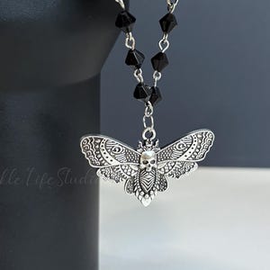 May include: A silver moth pendant with a skull design, hanging from a black beaded chain. The moth's wings display detailed patterns. The pendant is attached to a black object, possibly a mug handle. The background is a muted grey.