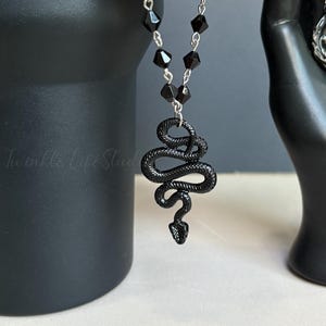 以下が含まれることがあります： シルバーチェーンと黒いビーズアクセントが付いた、黒いヘビのペンダントネックレス。ヘビのペンダントは複雑に詳細で、コイル状になっています。ネックレスは黒い手の像と黒いマグカップに飾られています。