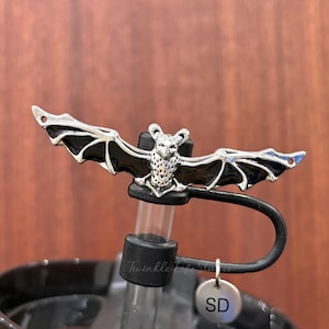 May include: A silver-colored bat-shaped bottle stopper with black wings, attached to a black straw. The stopper is on top of a black tumbler. A small, round charm with the letters "SD" hangs from the straw.