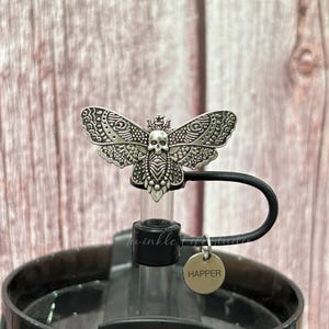 May include: A silver-colored bottle stopper featuring a detailed moth design with a skull motif. The stopper is attached to a black water bottle with a small tag that says "Happer".