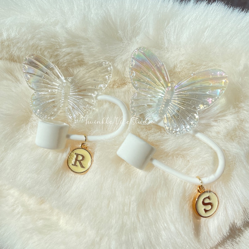 Puede incluir: Dos gomas para el pelo blancas con abalorios de mariposa iridiscente y abalorios de letras doradas. Los abalorios est&aacute;n unidos a las gomas para el pelo con una cadena dorada. Las gomas para el pelo est&aacute;n sobre una superficie blanca y esponjosa.