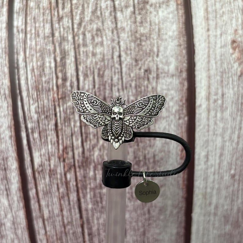 May include: A silver-colored moth-shaped bottle stopper with a skull design. The stopper has a black loop and a small, round charm that says "Sophia". The bottle stopper is on a clear tube. The background is a wooden surface.