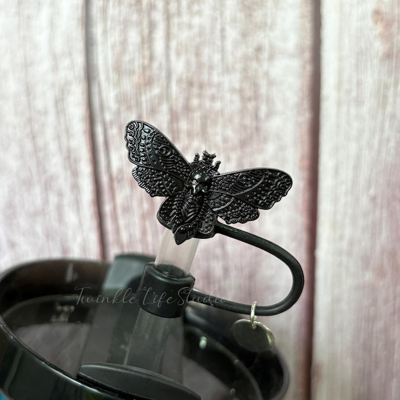 May include: Black metal moth-shaped lid topper. The topper is attached to a black lid with a curved handle and a small charm. The moth has detailed wing patterns and a crown. The lid is on a tumbler.
