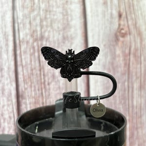 May include: Black butterfly-shaped tumbler topper with a straw and a small silver tag that says "Olivia". The topper is attached to a black tumbler. The background is a light brown wooden surface.