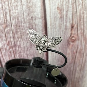 May include: A silver-colored bottle stopper featuring a moth design with a skull on its body. The moth has detailed wing patterns. The stopper is attached to a black lid, with the text "Twinkle Life" visible. A small, round tag hangs from the lid.