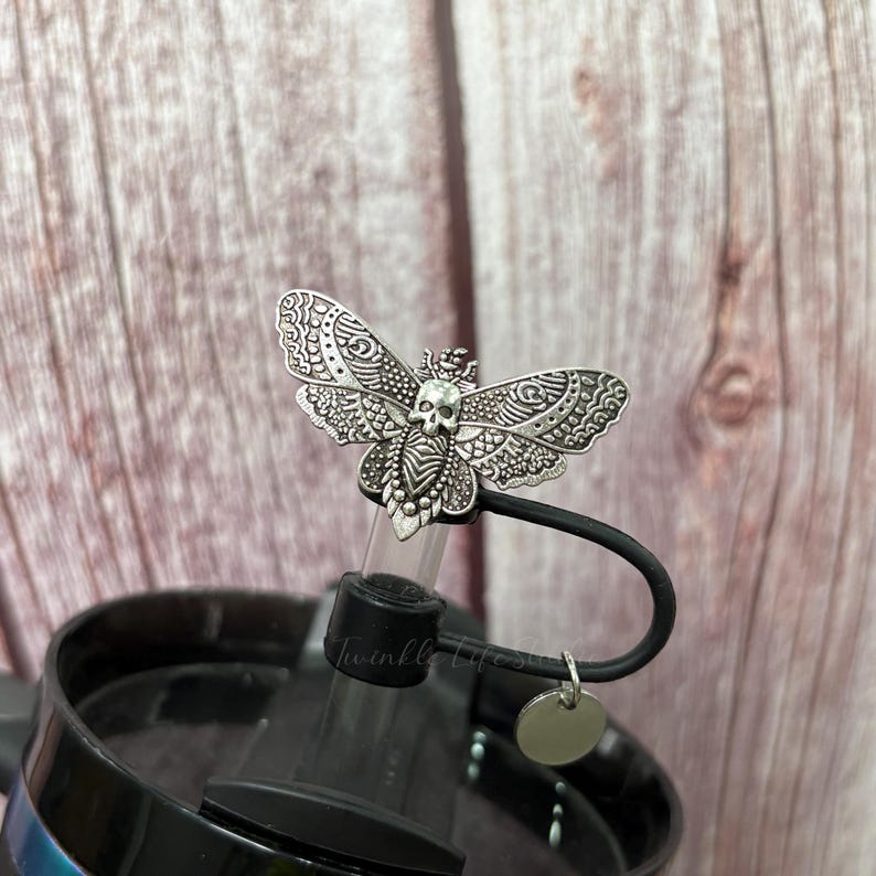May include: A silver-colored bottle stopper featuring a moth design with a skull on its body. The moth has detailed wing patterns. The stopper is attached to a black lid, with the text "Twinkle Life" visible. A small, round tag hangs from the lid.