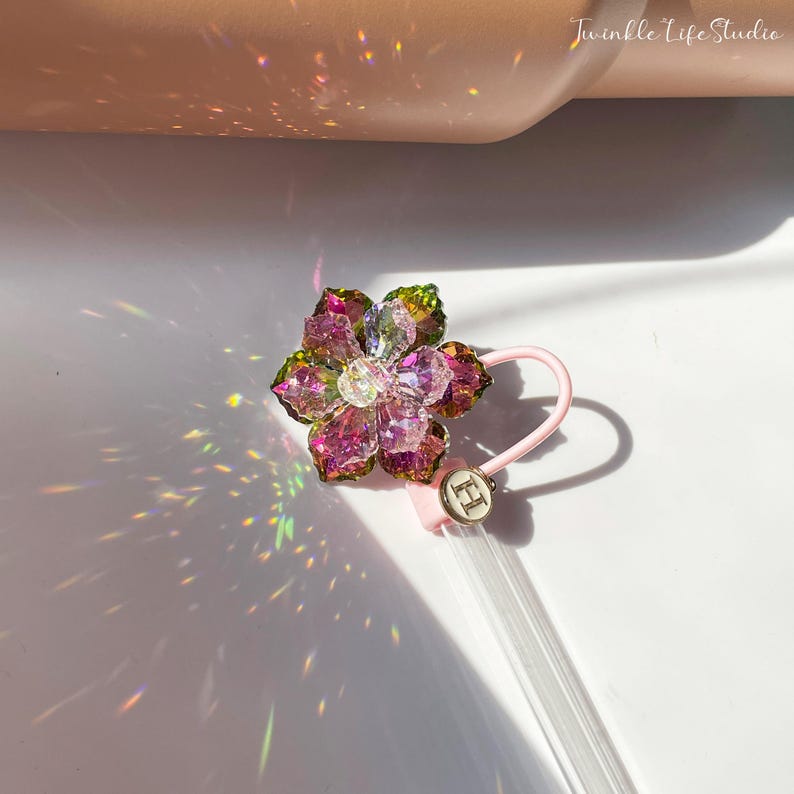 May include: A pink and green crystal flower charm with a clear crystal center. The charm is attached to a pink and white string with a silver charm that says "HI".
