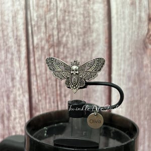 May include: A silver-colored moth-shaped bottle stopper with a skull design. The bottle stopper is attached to a black handle and a clear bottle. The text "Twinkle Life Studio" is visible on the bottle. A small, round tag with the name "Olivia" hangs from the bottle.